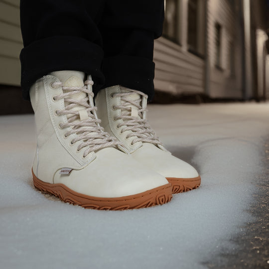 Off white barefoot winter boots worn while standing in the snow during an evening city scene, showcasing warmth, water resistance, and natural barefoot movement.