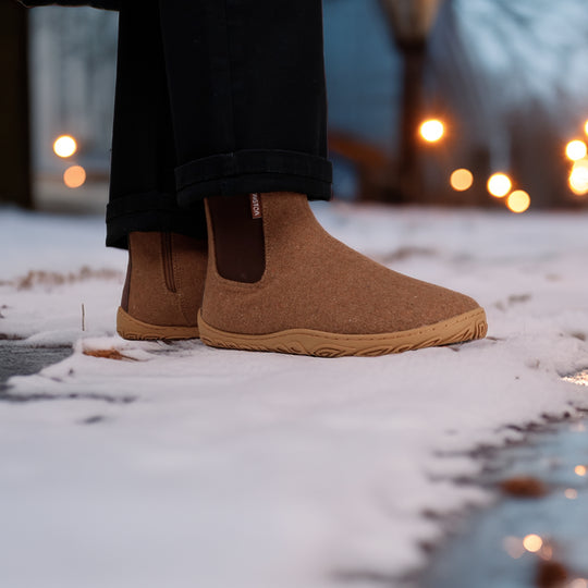Roasted almond barefoot winter felt Chelsea boots worn with black pants in a snowy winter setting, shown from the side to highlight their natural movement and warm design.