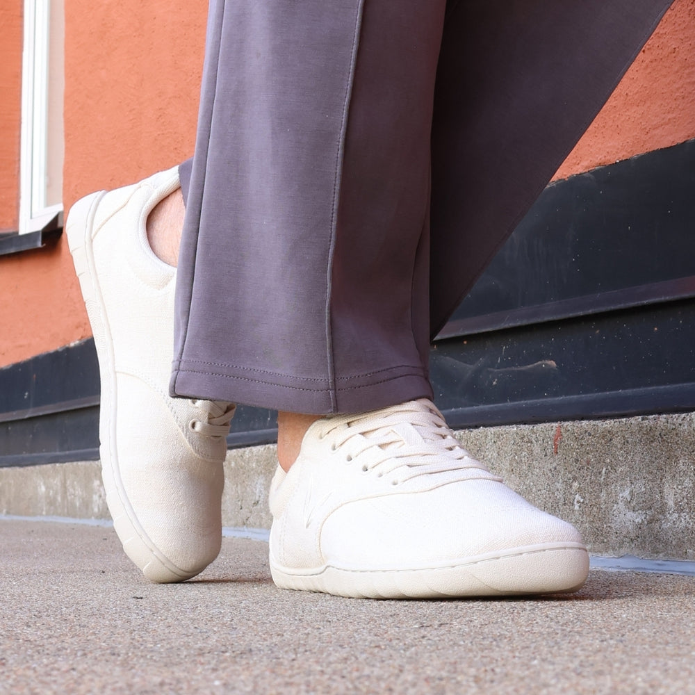 Vaistoa Huoleton barefoot hemp sneakers – natural hemp, shoes worn in the city with wide dark purple trousers, mid-step in front of a coloured wall