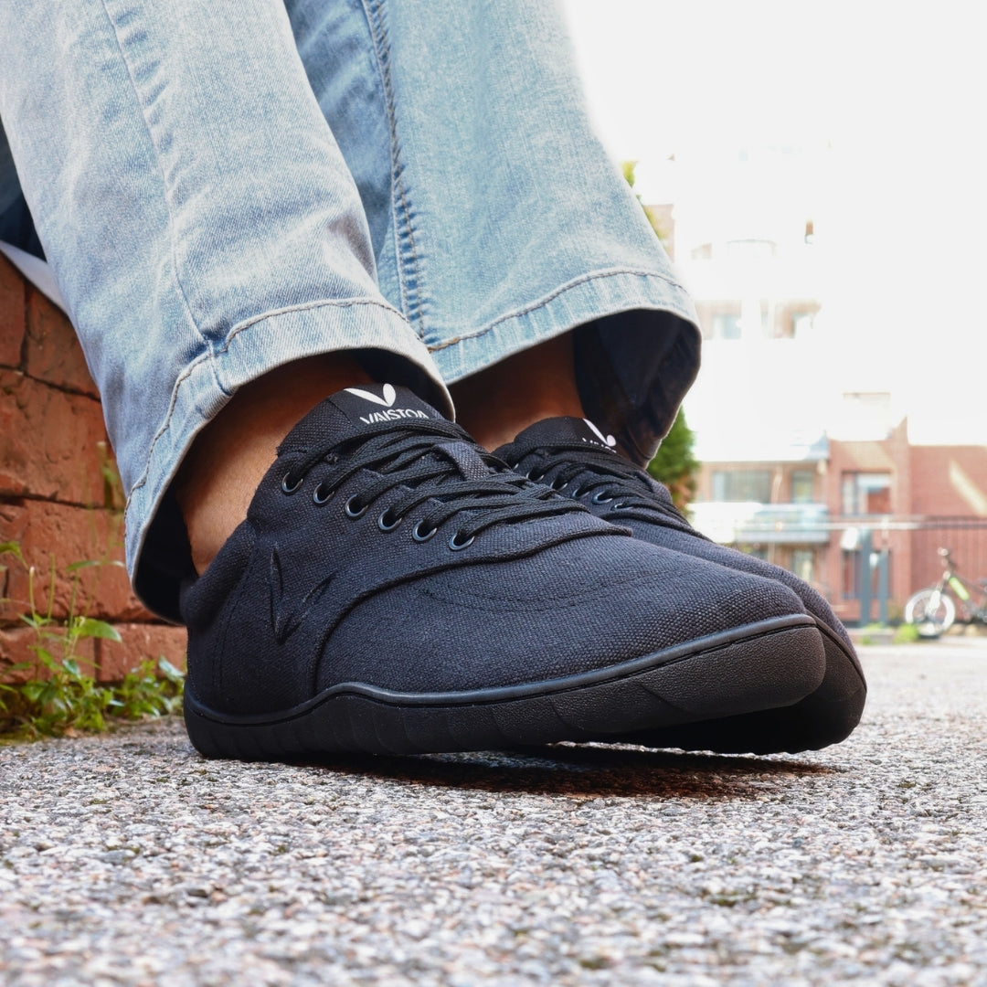 Vaistoa Huoleton barefoot hemp sneakers – soft black, shoes worn on a gravel path with light blue jeans, red brick wall in the background
