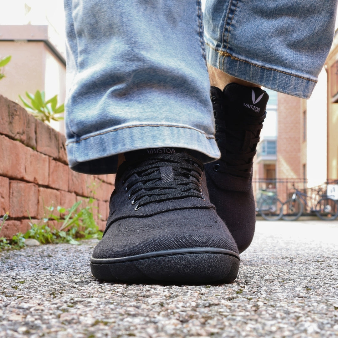 Vaistoa Huoleton barefoot hemp sneakers – soft black, shoes worn mid-step on a gravel path with light blue jeans, red brick wall in the background