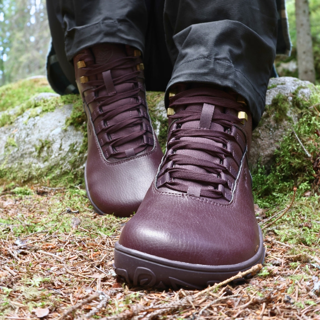 Vaistoa Uoma 2 barefoot trekking boots plum red – close-up of the boots being worn, the wearer's feet resting on pine-needle litter in front of a mossy boulder