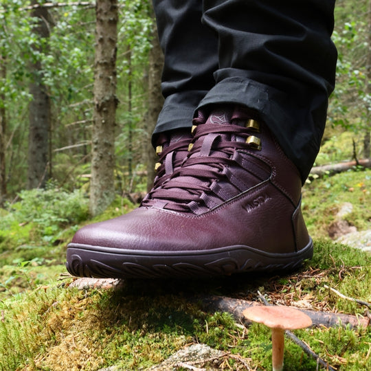 Vaistoa Uoma 2 barefoot trekking boots plum red – close-up of a single boot being worn, seen from the side, resting on a fallen branch among moss with a mushroom growing in the foreground