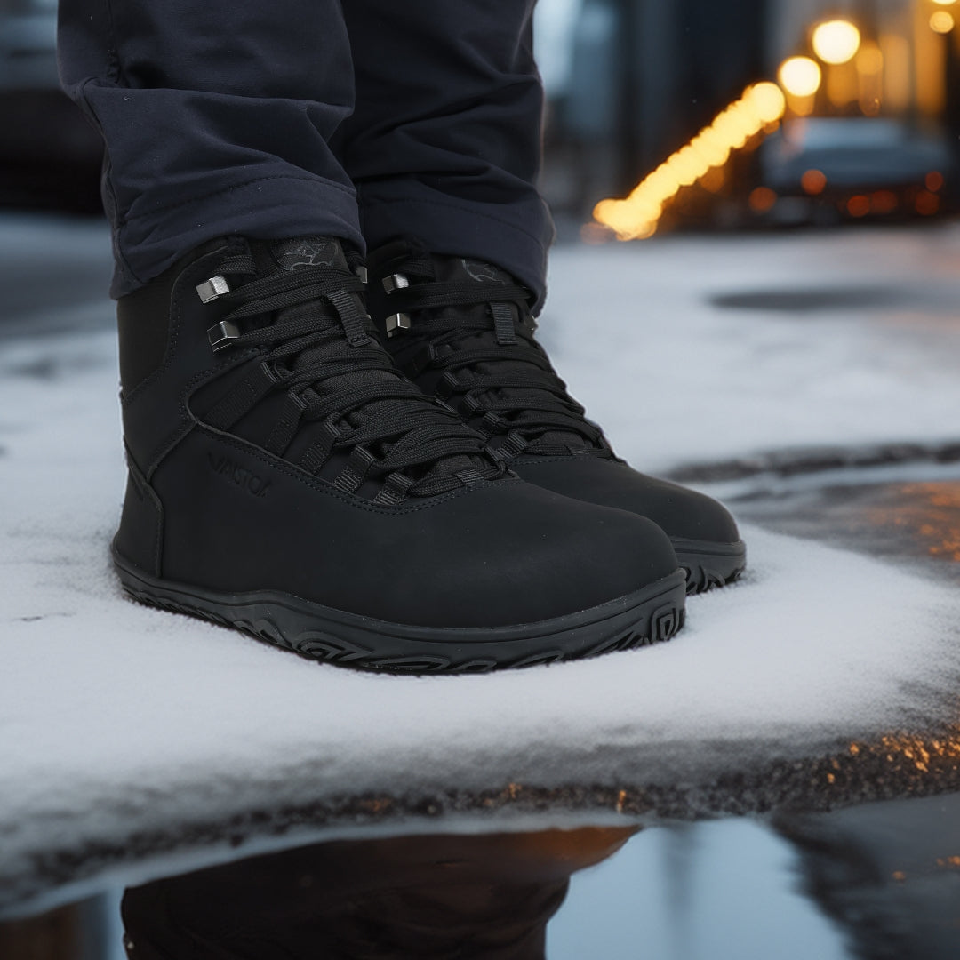 Käyttökuva noenmustista Vaistoa Uoma Talvi 3 -paljasjalkakengistä, joissa otetaan askel iltavalaistulla kadulla. Kuvassa korostuu kengän luonnollinen liike ja pitävä pohja.