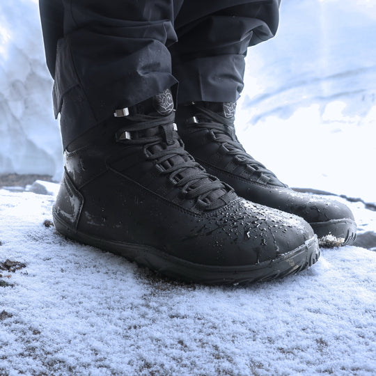 Black barefoot hiking boots shown from a front angle on a snowy rock in daylight, highlighting their waterproof design and winter grip sole.