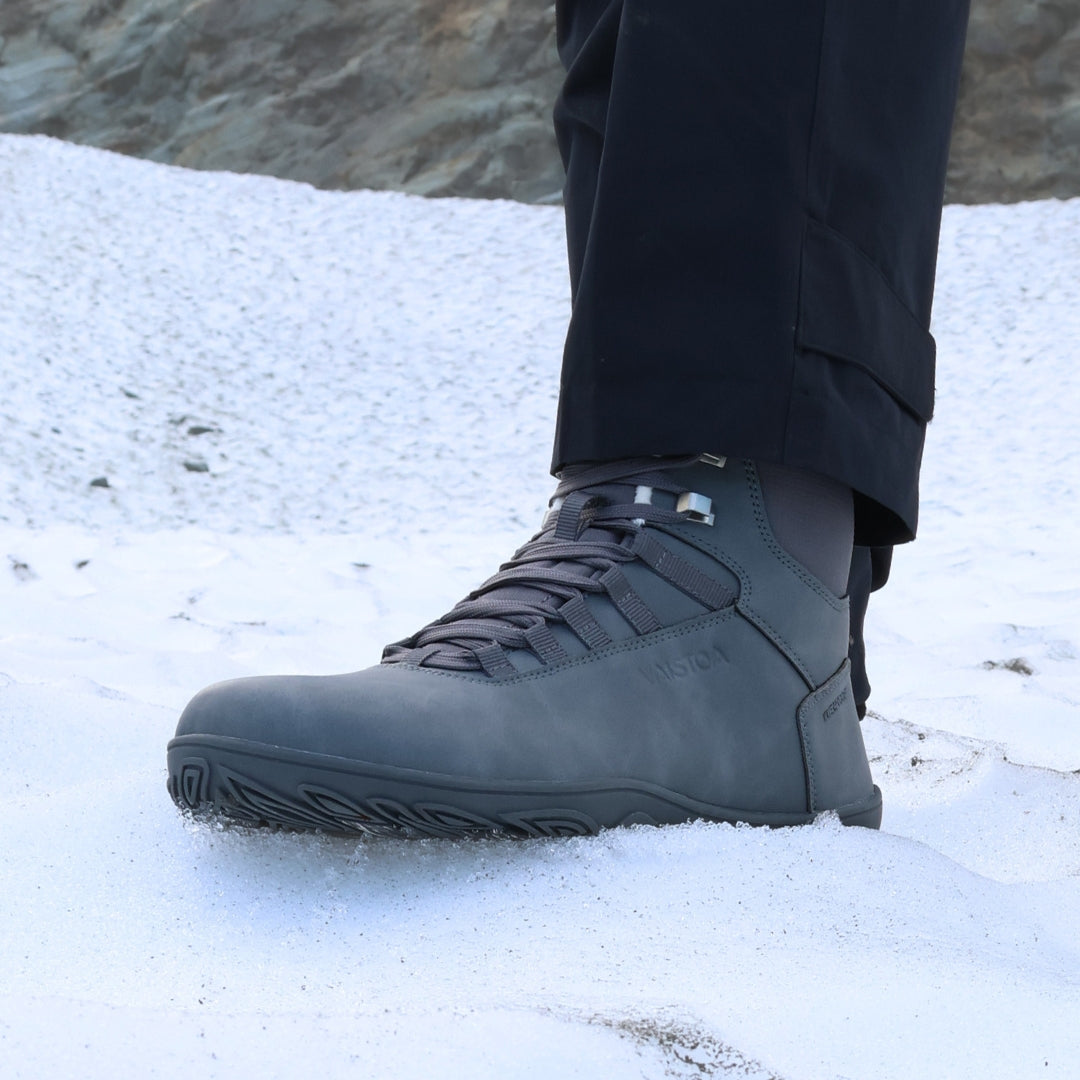 Frost gray barefoot hiking boots shown from a front angle on a snowy hill in daylight, emphasizing their waterproof design, winter grip sole, and natural barefoot comfort.