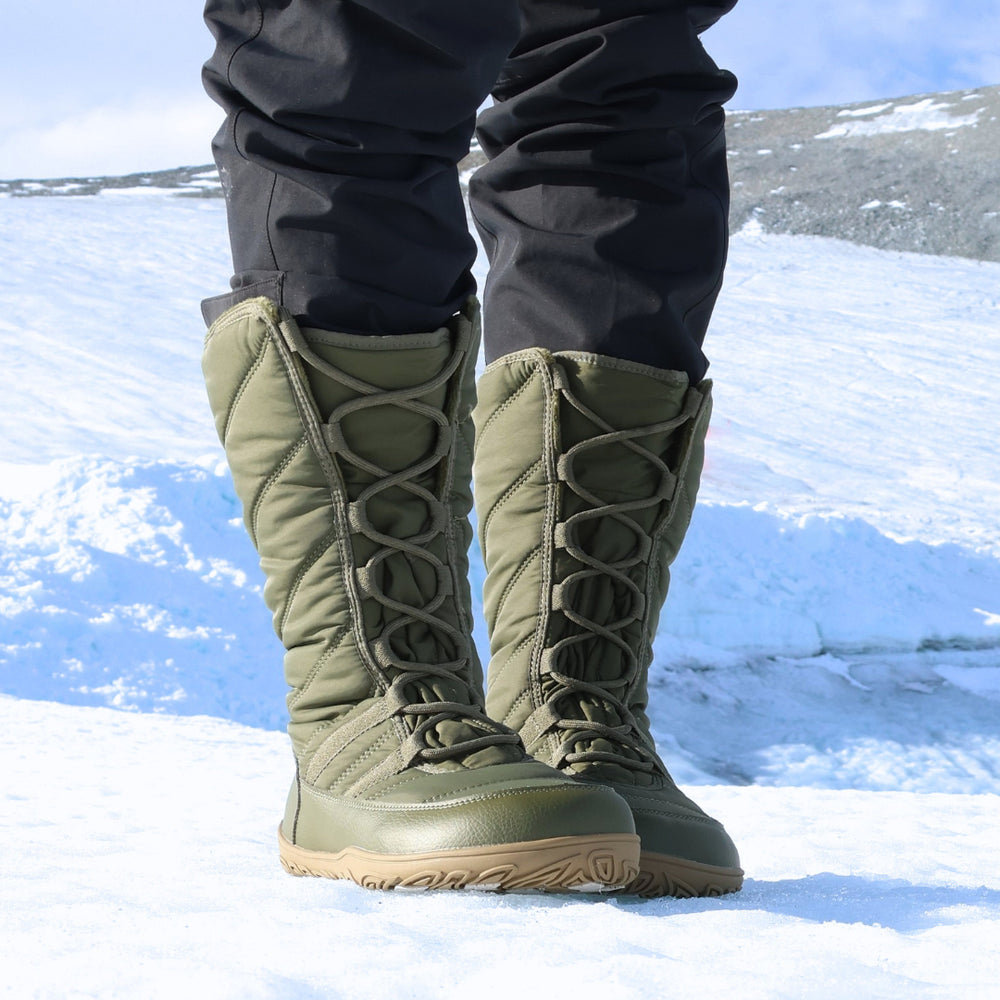 Vaistoa Hanki 3 barefoot winter snow boots, colour Olive green – lifestyle photo: pair from the front standing on snow with black trousers, lacing visible.