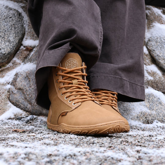 Vaistoa Hurmos 3 casual winter barefoot boots – lifestyle photo: pair from a front angle on a snowy rock with dark brown trousers, lacing and the Vaistoa logo on the tongue visible