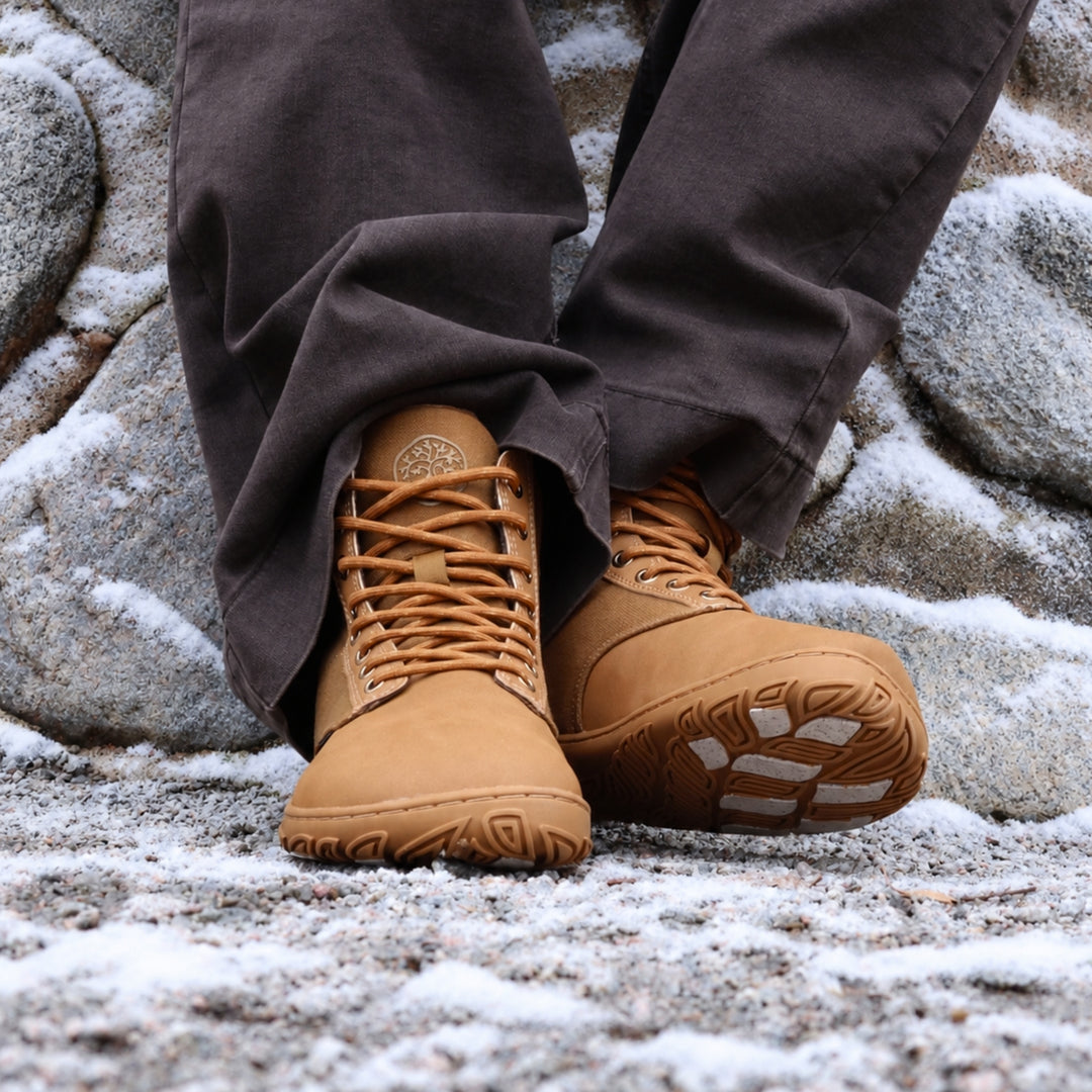 Vaistoa Hurmos 3 casual winter barefoot boots – lifestyle photo: pair on a snowy rock with dark brown trousers, one shoe tilted so the grip pads on the outsole are visible, a stone wall in the background
