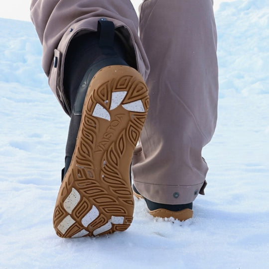 Vaistoa Hurmos 3 casual winter barefoot boots – lifestyle photo: a step in the snow from behind with taupe trousers, the honey-brown outsole and the light grip pads visible