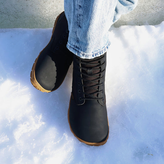 Vaistoa Hurmos 3 casual winter barefoot boots – lifestyle photo: legs crossed, shot from above in sunny snow with blue jeans, lacing and the honey-brown edge of the outsole visible