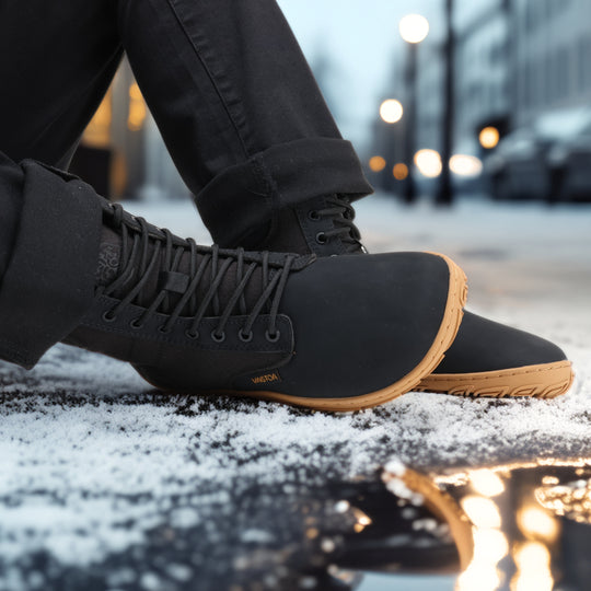 Vaistoa Hurmos 3 casual winter barefoot boots – lifestyle photo: pair from the side while seated on a snowy street in the evening with black trousers, city lights and a puddle reflection in the background