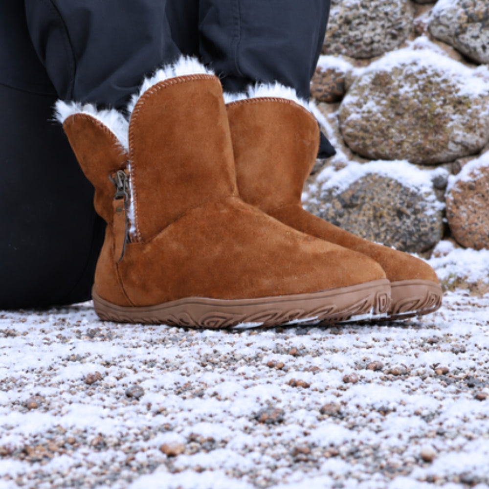 Vaistoa Huurre 2 barefoot winter ankle boots, colour Caramel brown – lifestyle photo: boots worn on snowy ground, black trousers, sitting against a stone wall.