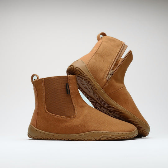 Vaistoa Kajo 2 barefoot chelsea boots – almond brown pair with the front shoe on its side showing the elastic panel and the rear shoe tilted so the leaf-patterned outsole is fully visible against a light background