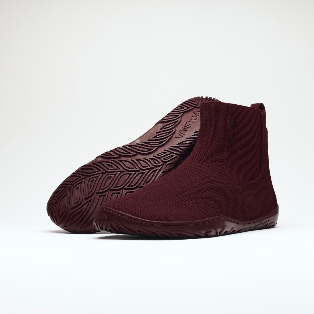 Vaistoa Kajo 2 barefoot chelsea boots – burgundy pair with the front shoe on its side showing the elastic panel and the rear shoe tilted so the leaf-patterned outsole is fully visible against a light background