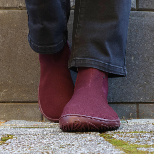 Vaistoa Kajo 2 barefoot chelsea boots – burgundy shoes worn on foot on mossy stone slabs in front of a concrete wall, black jeans