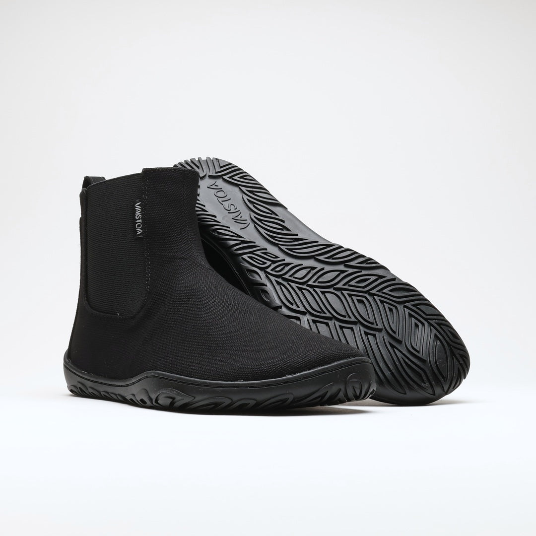 Vaistoa Kajo 2 barefoot chelsea boots – classic black pair with the front shoe on its side showing the elastic panel and the rear shoe tilted so the leaf-patterned outsole is fully visible against a light background