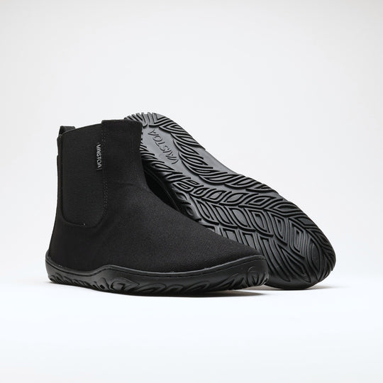 Vaistoa Kajo 2 barefoot chelsea boots – classic black pair with the front shoe on its side showing the elastic panel and the rear shoe tilted so the leaf-patterned outsole is fully visible against a light background
