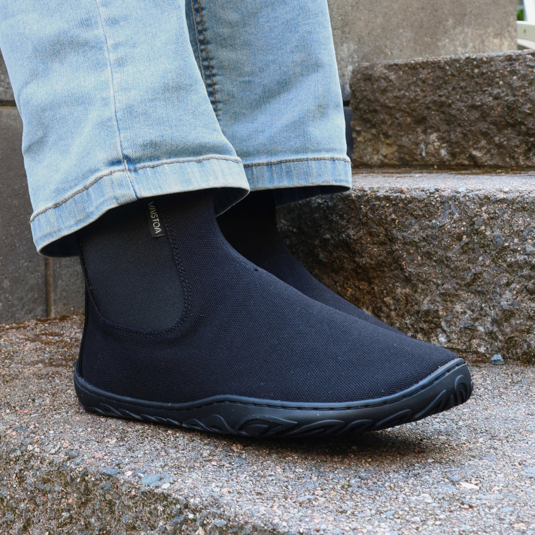 Vaistoa Kajo 2 barefoot chelsea boots – classic black shoe worn on foot on a granite step shown from the side, elastic panel and Vaistoa label visible, light jeans