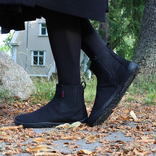 Vaistoa Kajo barefoot Chelsea boots – black, boots worn with dark tights on a leaf-covered path, mid-step so the outsole is visible, a house in the background