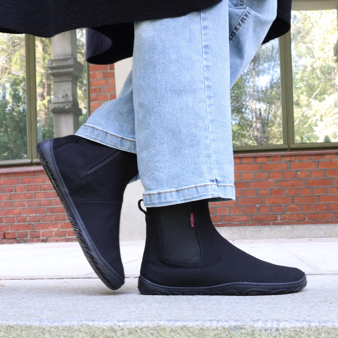 Vaistoa Kajo barefoot Chelsea boots – black, boots worn with light blue jeans, standing on a pavement in front of a red brick wall and a window