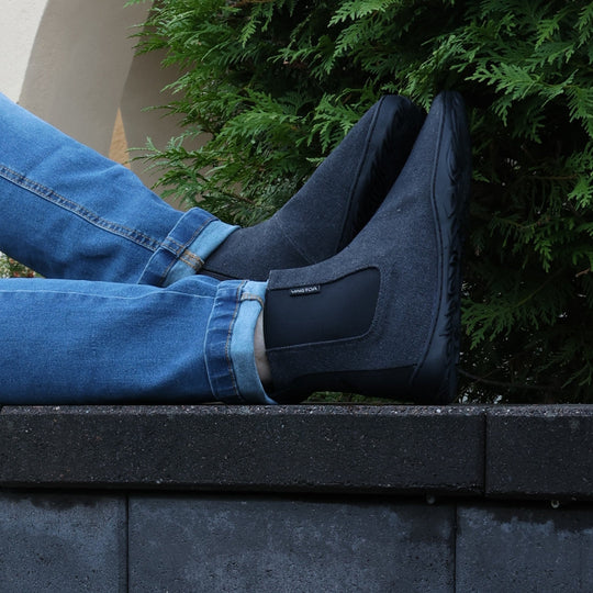 Vaistoa Kajo barefoot Chelsea boots – smoky stonewashed grey, boots worn with blue jeans, seated with one foot raised on a stone ledge, conifers in the background