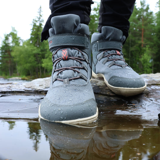 Vaistoa Kaste waterproof barefoot shoes for kids blue grey – worn shoes standing in a shallow puddle on wet rock, water droplets on the upper and a spruce forest behind