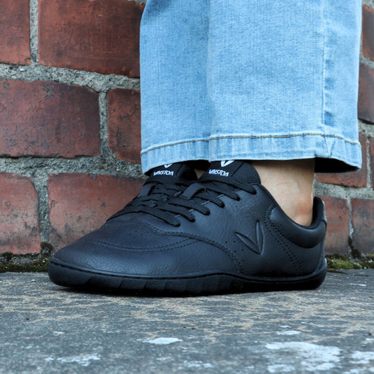 Vaistoa Katu 2 black barefoot sneakers in use – person standing outdoors in blue jeans on an autumn stone surface, pictured from the front.