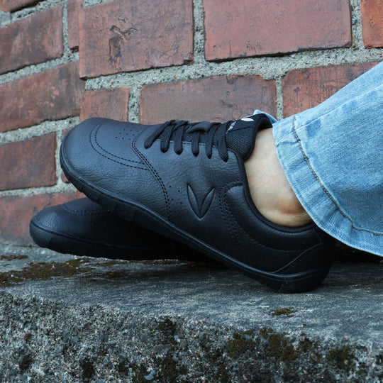 Vaistoa Katu 2 black barefoot sneakers in use – person sitting outdoors on an autumn day in blue jeans, feet up against a brick wall.