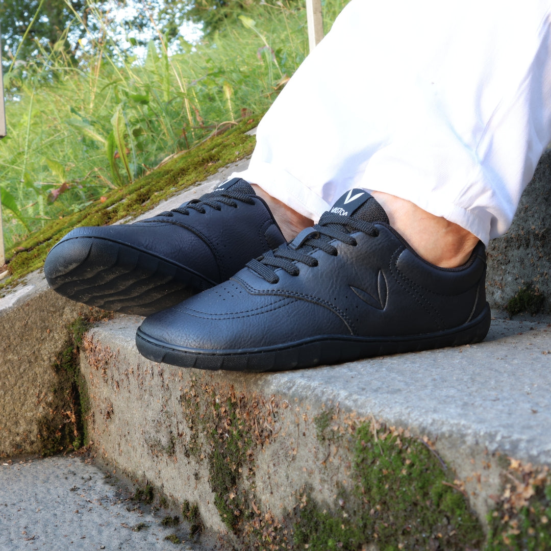 Vaistoa Katu 2 black barefoot sneakers in use – person sitting outdoors on stairs on a summer day, wearing white jeans, pictured close-up.