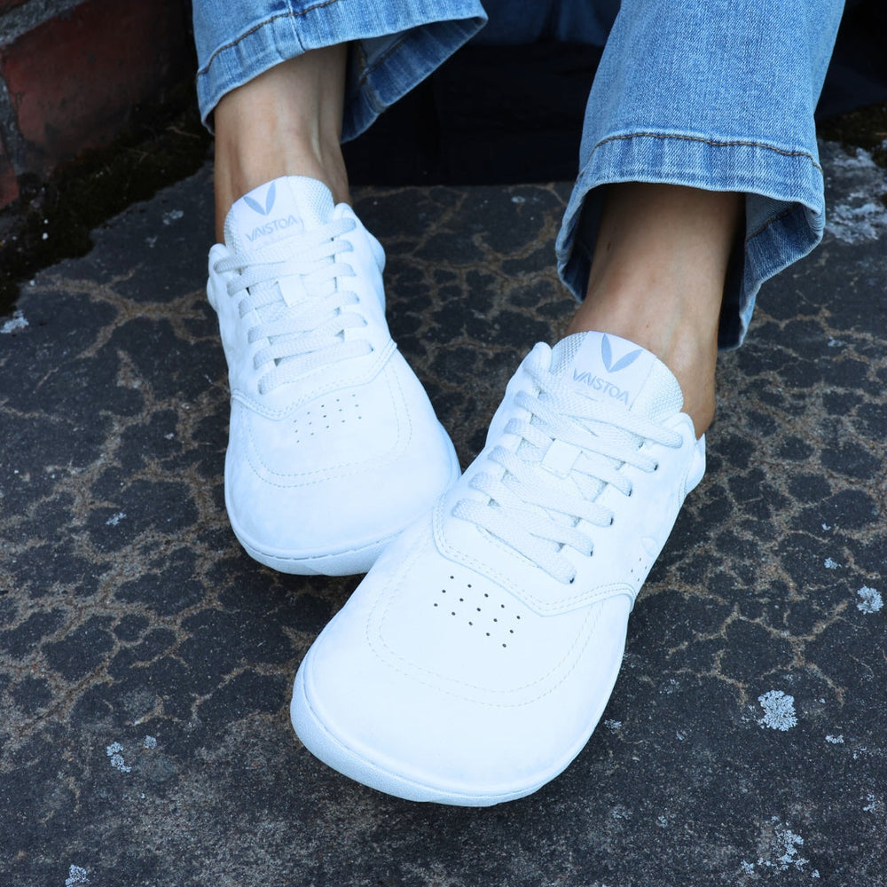 Vaistoa Katu 2 white barefoot sneakers in use – person sitting outdoors in blue jeans on an autumn stone surface, pictured from the front.