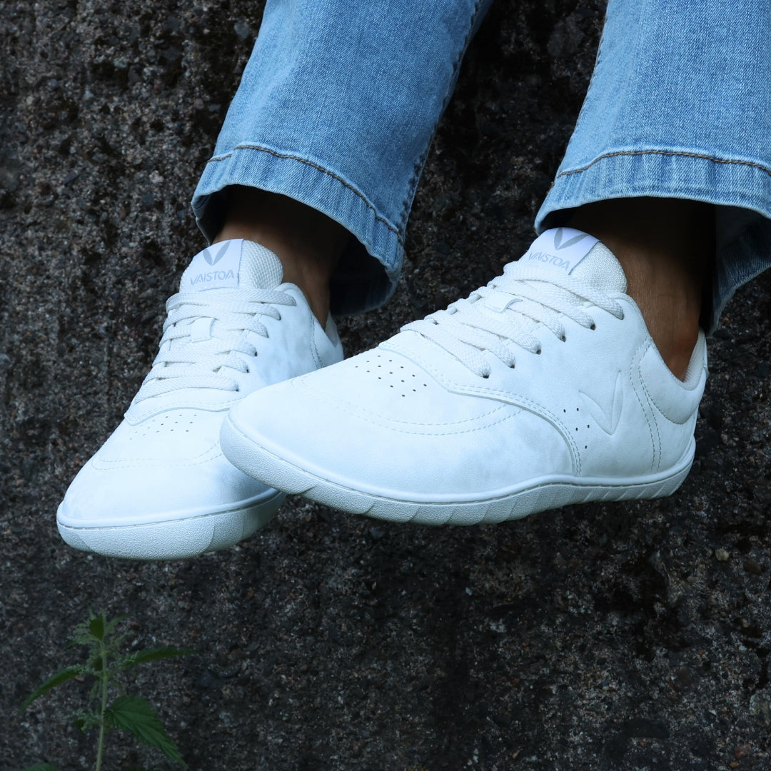 Vaistoa Katu 2 white barefoot sneakers in use – person sitting outdoors on an autumn day in blue jeans, feet up against a dark-textured wall.
