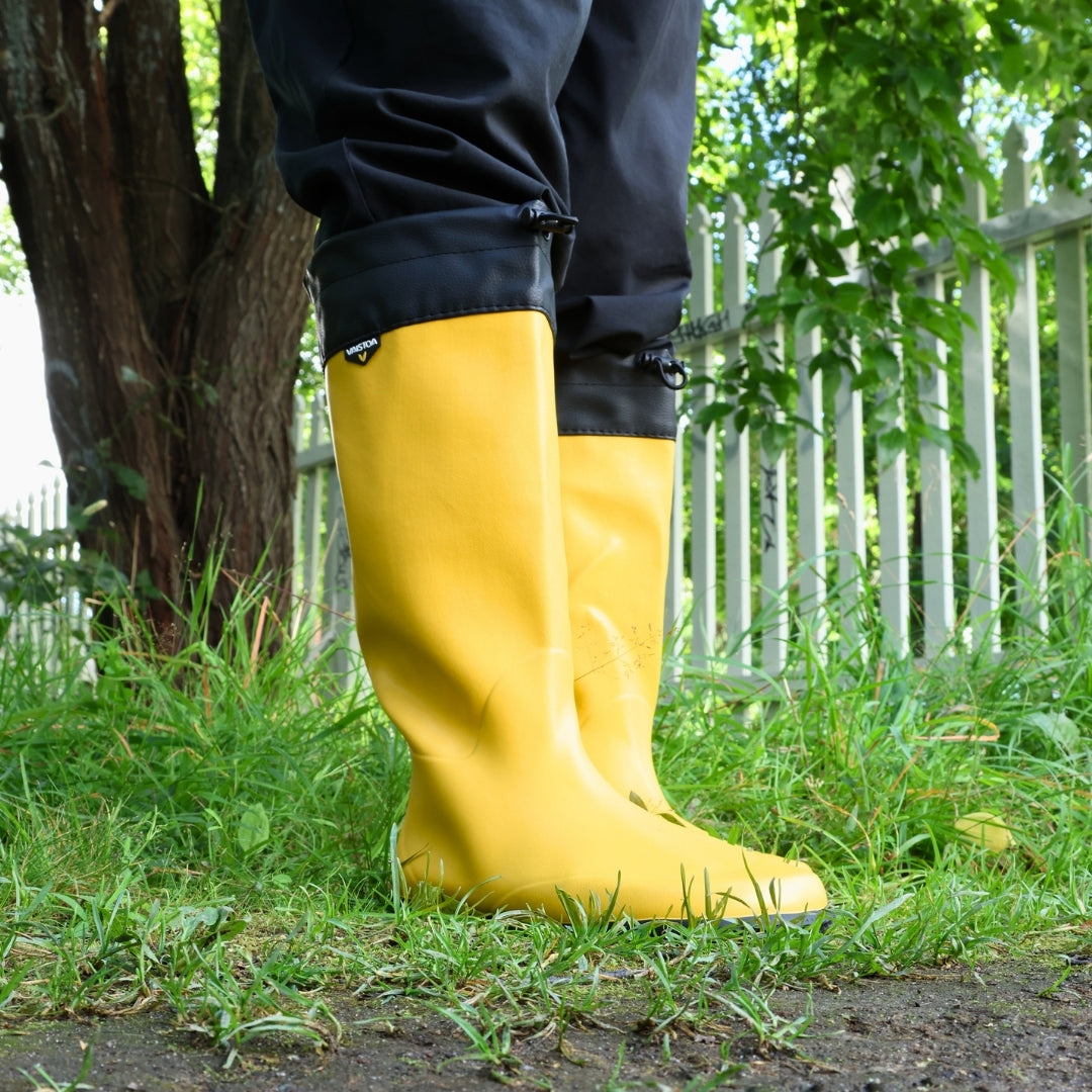 Vaistoa Katve 2 barefoot rubber boots yellow worn on the grass beside a tree and a white picket fence