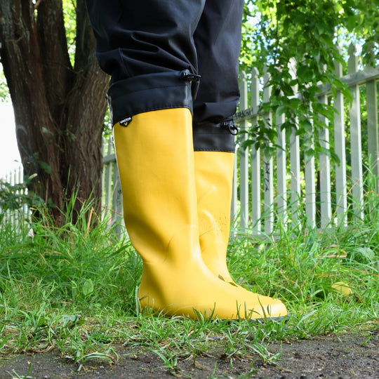 Vaistoa Katve 2 barefoot rubber boots yellow worn on the grass beside a tree and a white picket fence
