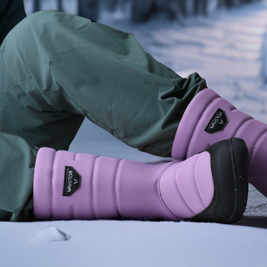 Vaistoa Kinos – lasten korkeavartiset paljasjalkakengät talveen, väri lila. Käyttökuva: edestä kuvattuna, lapsi istuu lumella siniset housut jalassa.