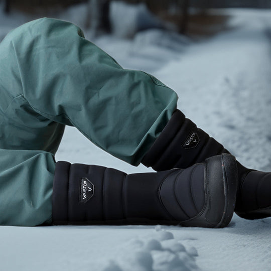 Vaistoa Kinos – lasten korkeavartiset paljasjalkakengät talveen, väri musta. Käyttökuva: edestä kuvattuna, lapsi istuu lumella siniset housut jalassa.