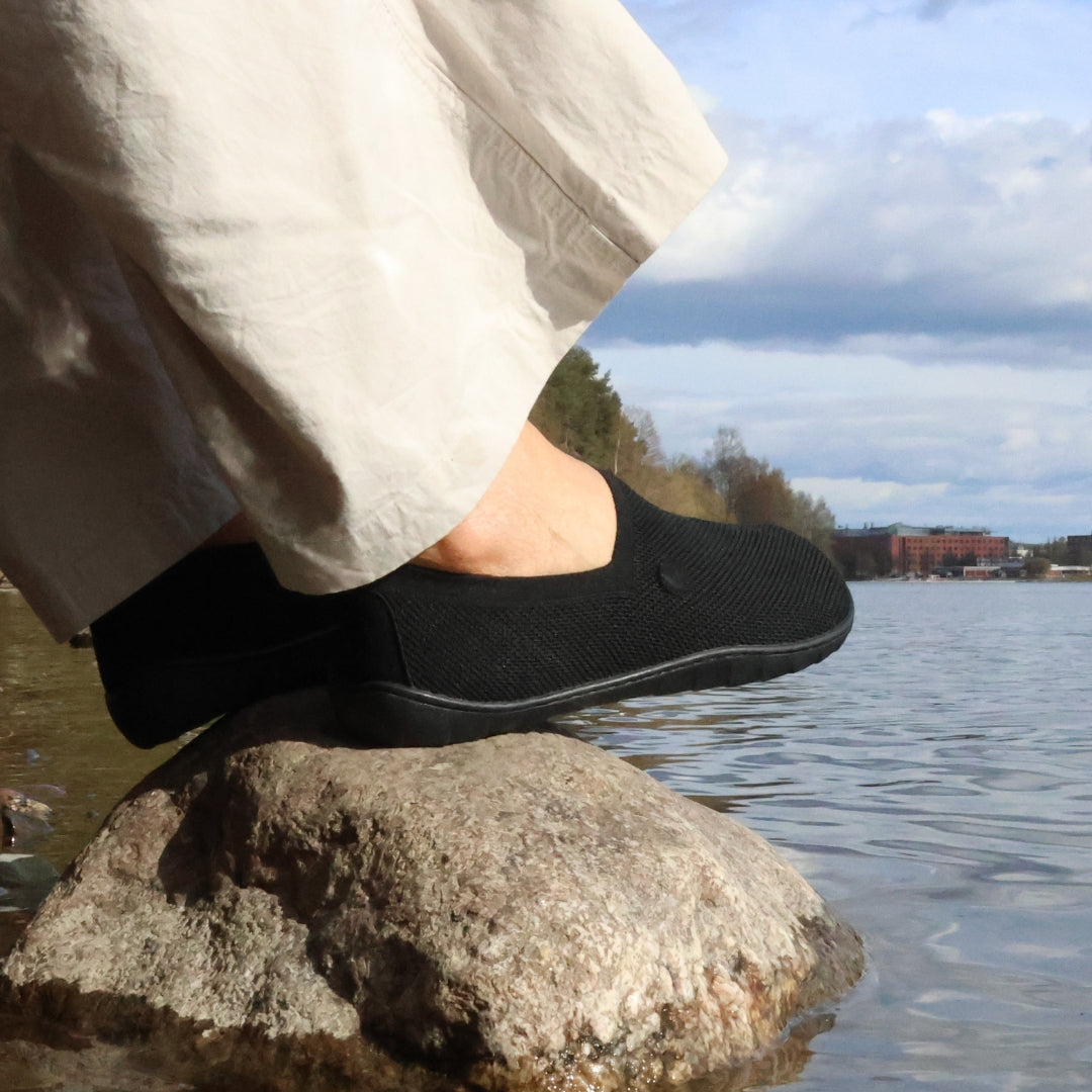 Vaistoa Lempeä 2 barefoot slip-on shoes black – lifestyle photo, beige pants sitting by the lake