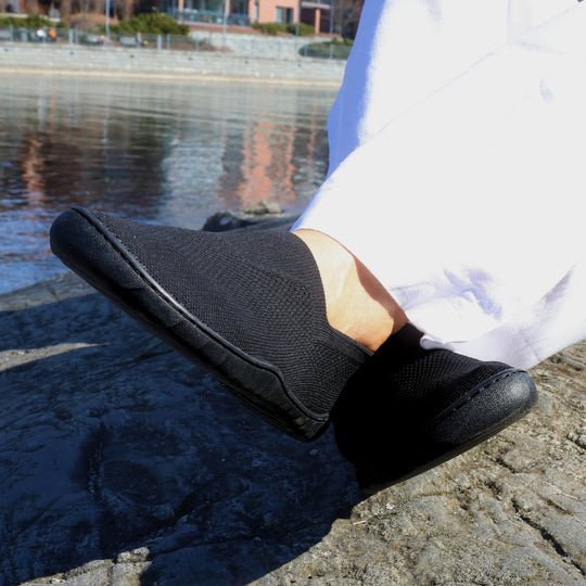 Vaistoa Lempeä 3 slip-on barefoot shoes – black shoes worn on feet resting crossed on a rock by the water