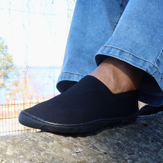Vaistoa Lempeä 3 slip-on barefoot shoes – black shoe worn on foot on a rock, side view with metal fence and water in the background