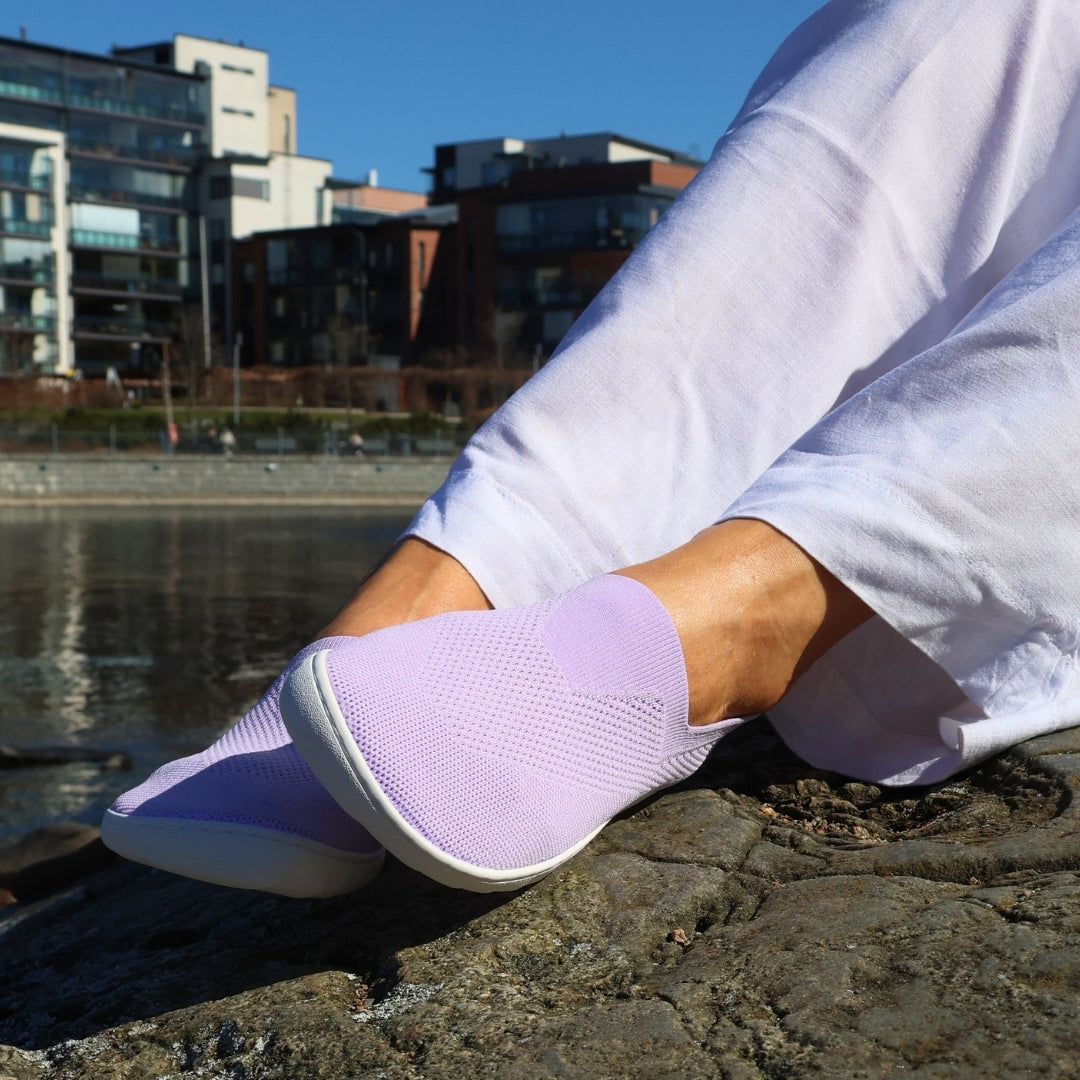 Vaistoa Lempeä 3 slip-on barefoot shoes – lavender shoes worn on feet from the side, resting on a rock with buildings and water in the background