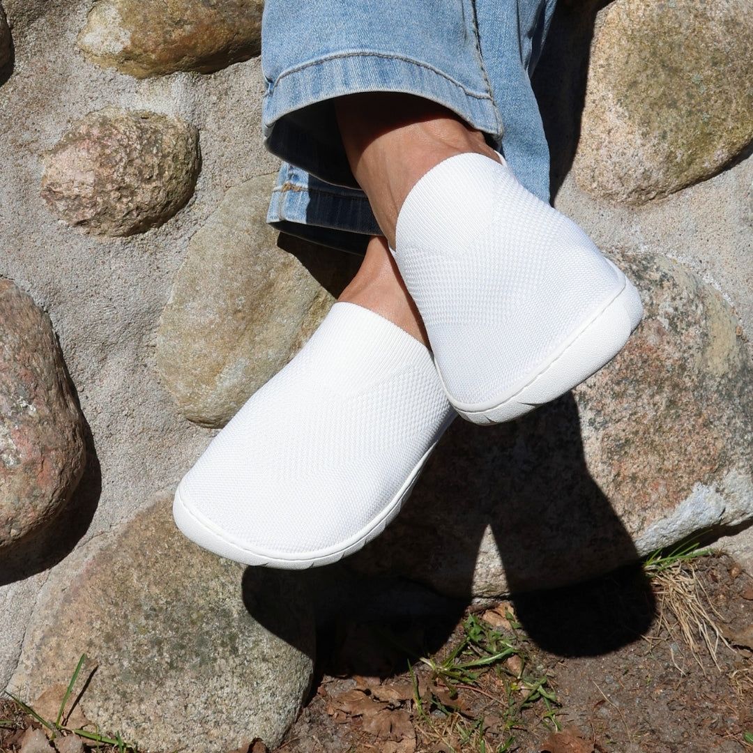 White Vaistoa Lempeä 3 slip-on barefoot shoes worn on feet against a stone wall outdoors