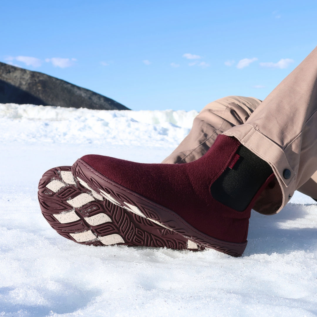 Vaistoa Sulo 2 barfota chelsea-boots för vinter – livsstilsbild: paret används sittande i snön med beige byxor, yttersulorna och de ljusa greppdynorna syns, ett fjäll i bakgrunden