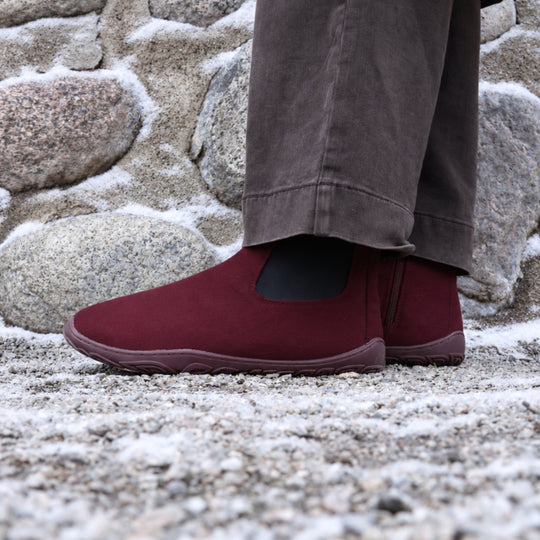 Vaistoa Sulo 2 barfota chelsea-boots för vinter – livsstilsbild: paret snett framifrån på en snöig klippa med mörkbruna byxor, den elastiska resåren syns, en stenmur i bakgrunden