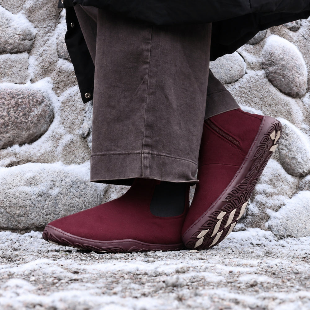Vaistoa Sulo 2 barfota chelsea-boots för vinter – livsstilsbild: paret på en snöig klippa med mörkbruna byxor, ena skon lutad så att greppdynorna på yttersulan syns, en stenmur i bakgrunden