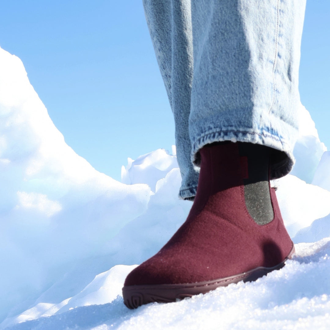 Vaistoa Sulo 2 barfota chelsea-boots för vinter – livsstilsbild: ett steg i snön snett framifrån med blå jeans, den elastiska resåren syns, en snövall och blå himmel i bakgrunden