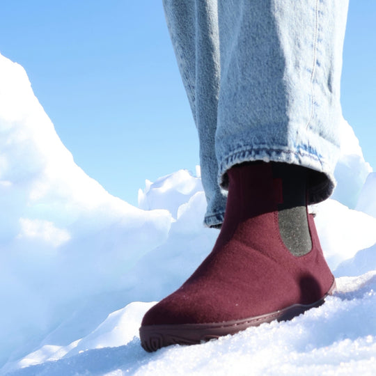 Vaistoa Sulo 2 barfota chelsea-boots för vinter – livsstilsbild: ett steg i snön snett framifrån med blå jeans, den elastiska resåren syns, en snövall och blå himmel i bakgrunden