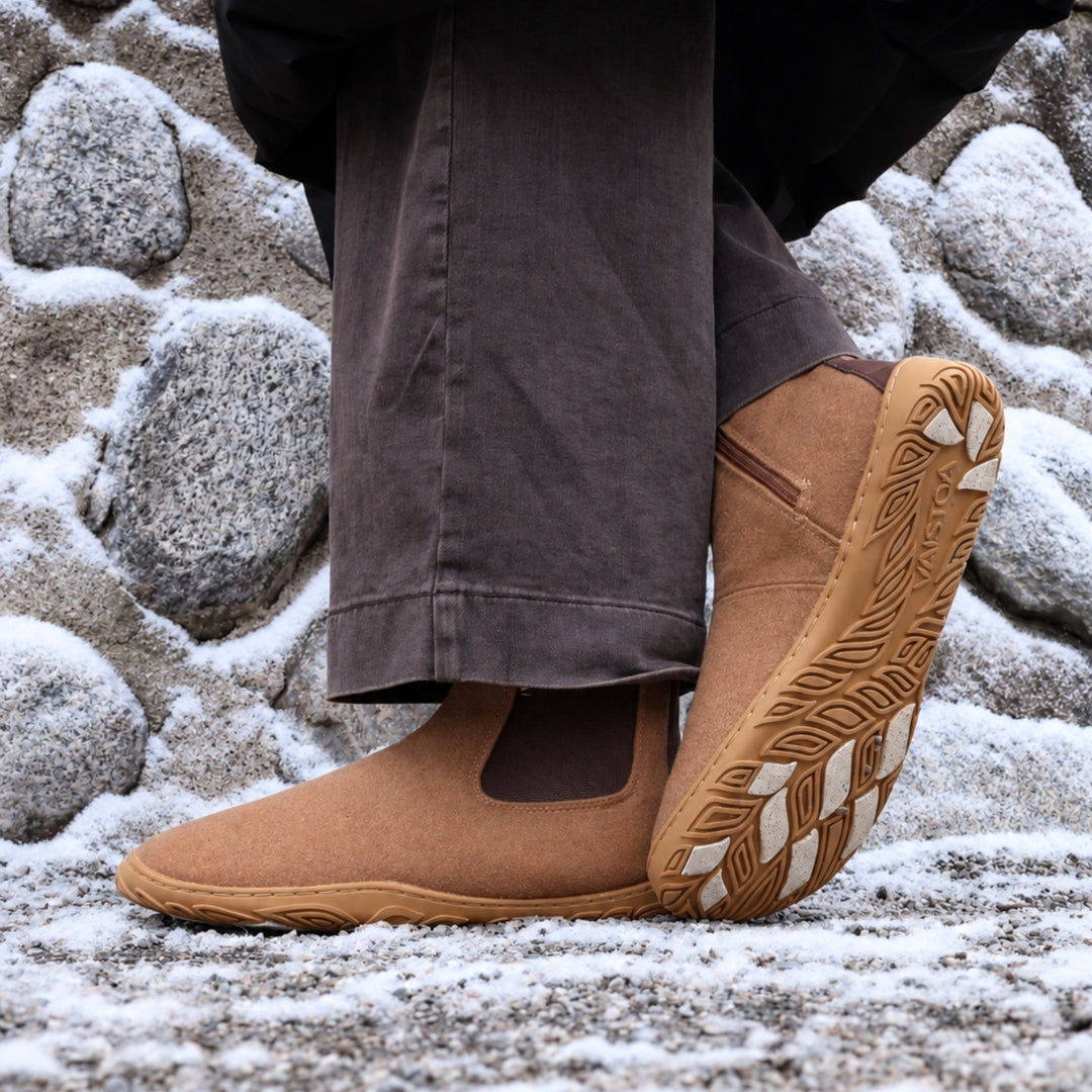 Vaistoa Sulo 2 barfota chelsea-boots för vinter – livsstilsbild: paret på en snöig klippa med mörkbruna byxor, ena skon lutad så att greppdynorna på yttersulan syns, en stenmur i bakgrunden