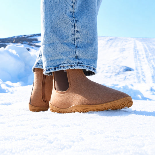 Vaistoa Sulo 2 barfota chelsea-boots för vinter – livsstilsbild: paret från sidan på snön med blå jeans, den elastiska resåren syns, blå himmel i bakgrunden