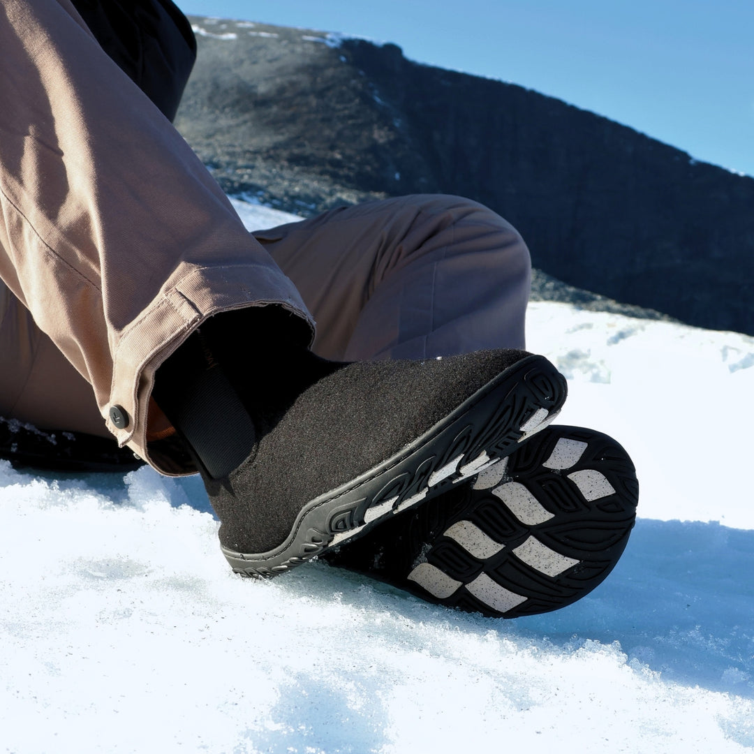 Vaistoa Sulo 2 barfota chelsea-boots för vinter – livsstilsbild: paret används sittande i snön med bruna byxor, yttersulan och de ljusa greppdynorna på ena skon syns, ett fjäll och blå himmel i bakgrunden