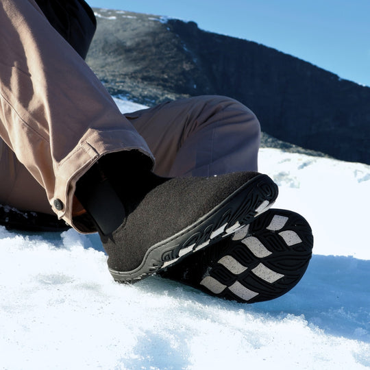 Vaistoa Sulo 2 barfota chelsea-boots för vinter – livsstilsbild: paret används sittande i snön med bruna byxor, yttersulan och de ljusa greppdynorna på ena skon syns, ett fjäll och blå himmel i bakgrunden